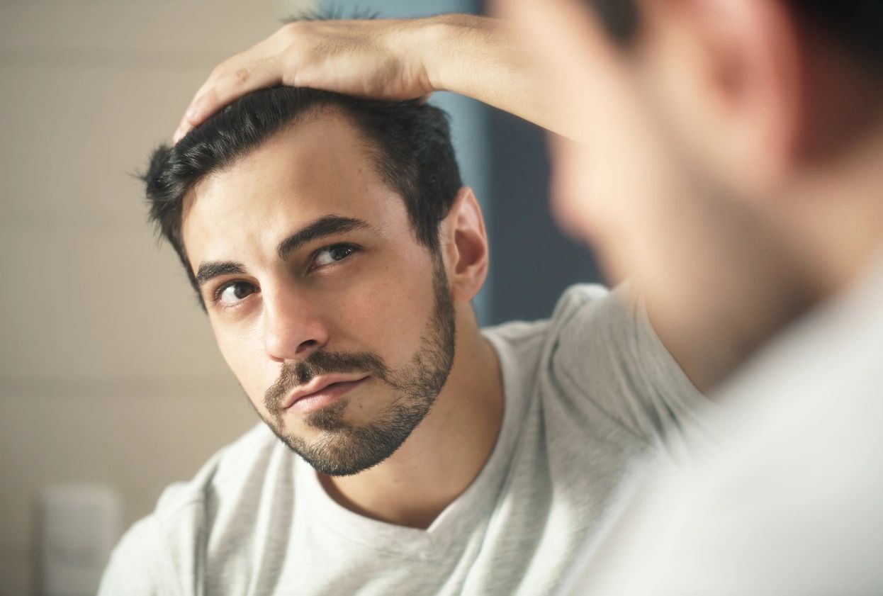 Man examining his hair in a bathroom mirror, touching the top of his head with a contemplative expression.