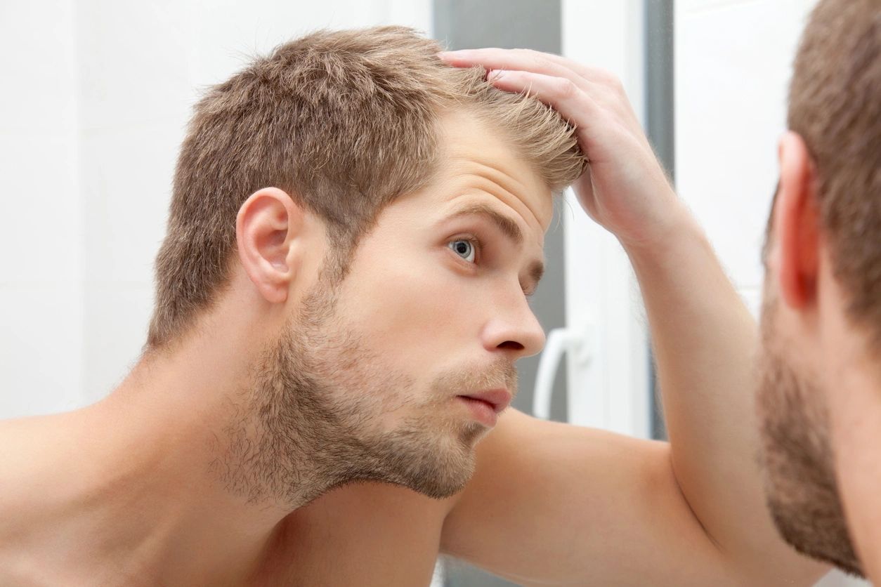 Man examining his hair in the mirror with a focused expression in a bathroom setting.